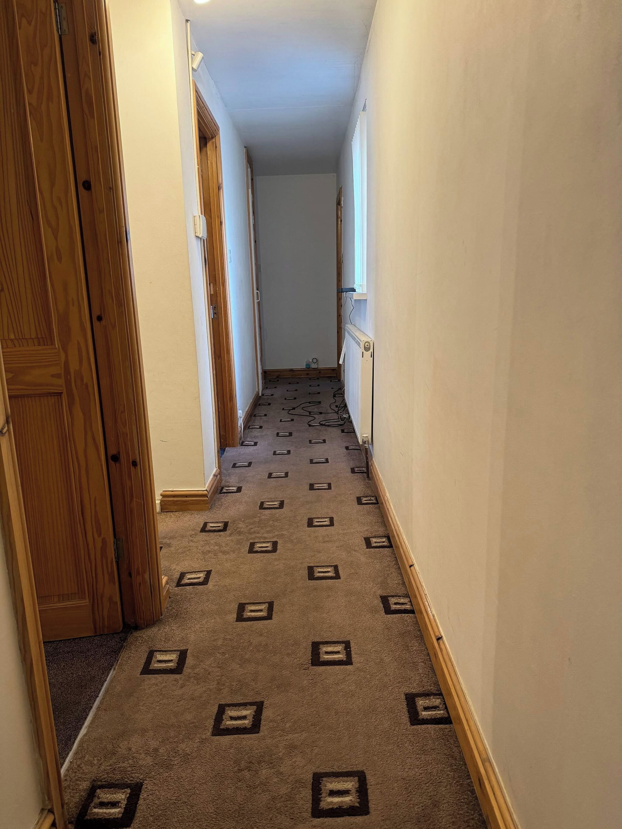A narrow hallway with patterned carpet, wooden doors, and a radiator on the wall.