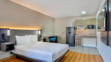 Quarto de hotel com cama, mesas de cabeceira, escrivaninha, cadeira, frigorífico, micro-ondas e cozinha compacta.