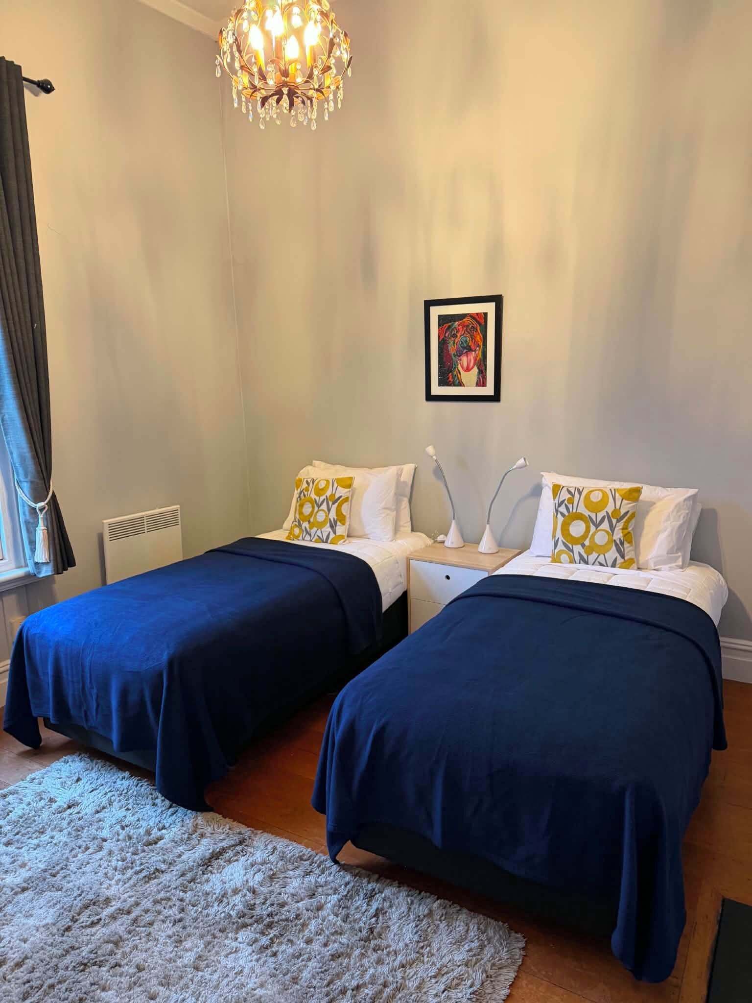Une chambre d’hôtel avec deux lits, un lustre, un tableau encadré au mur et une fenêtre avec des rideaux.