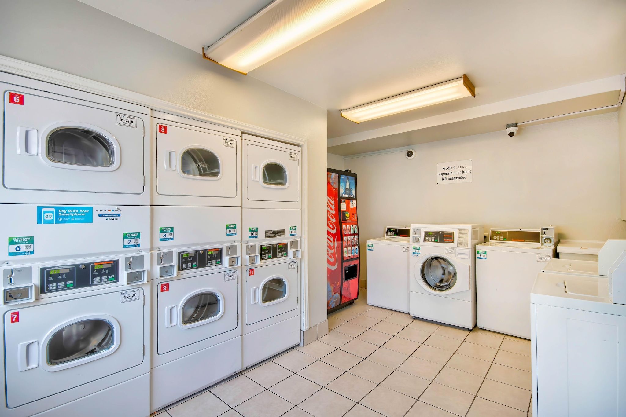 A laundry facility with multiple washing machines and dryers, a Coca-Cola vending machine, and a sign indicating Studio 6 is not responsible for unattended items.