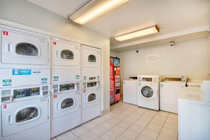 Et vaskeri med flere vaskemaskiner og tørretumblere, en Coca-Cola automat og et skilt, der angiver at Studio 6 ikke er ansvarlig for uovervågede genstande.