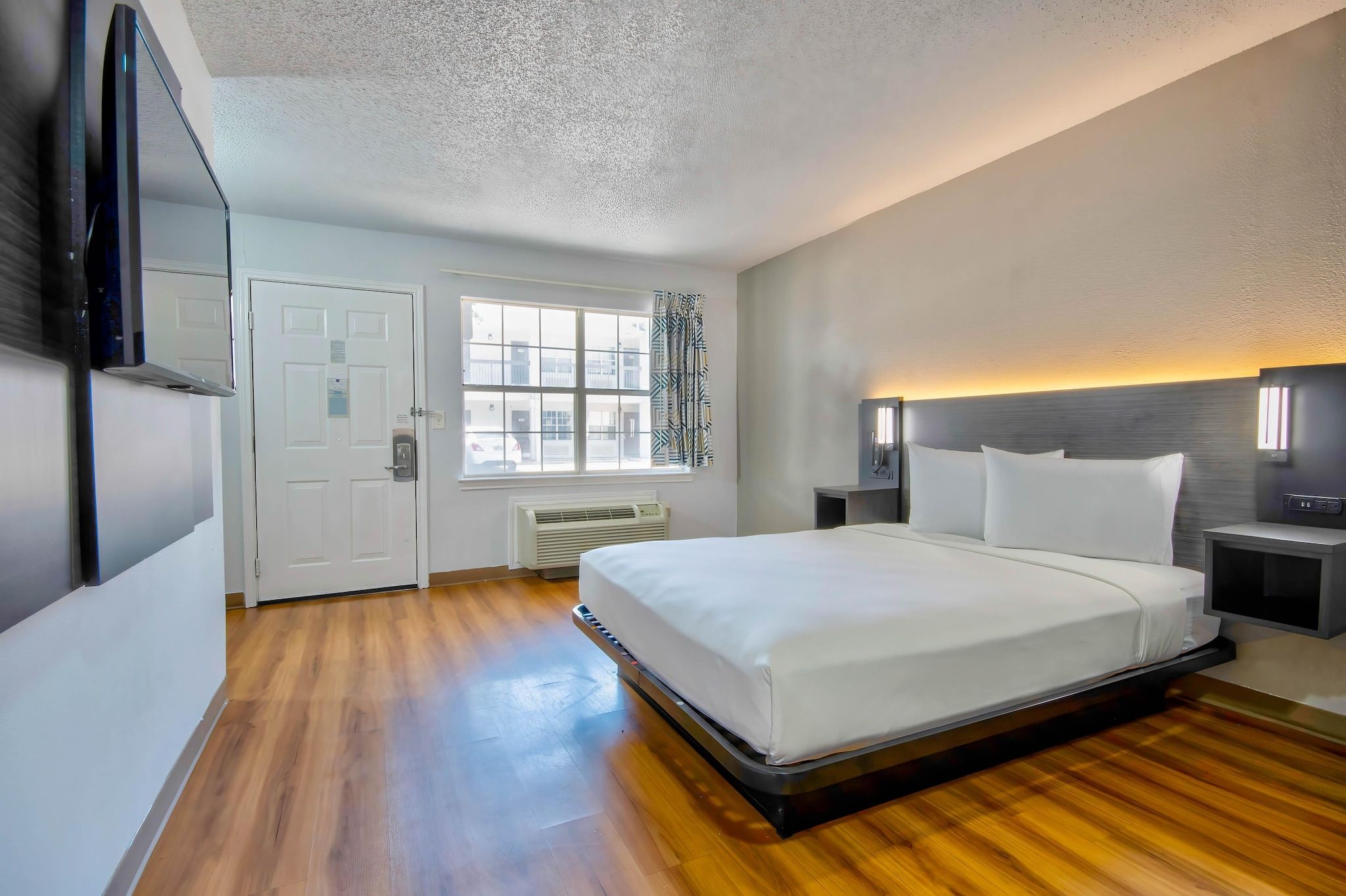 Quarto de hotel com uma cama grande, mesa de cabeceira, janela com cortinas, televisão na parede e ventilador de teto.