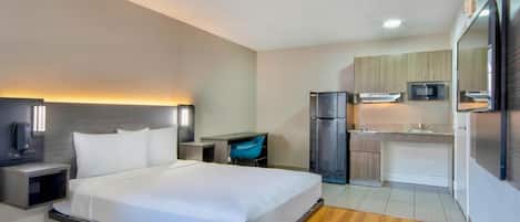 A hotel room with a bed, bedside table, lamp, and a kitchenette with a refrigerator and microwave.