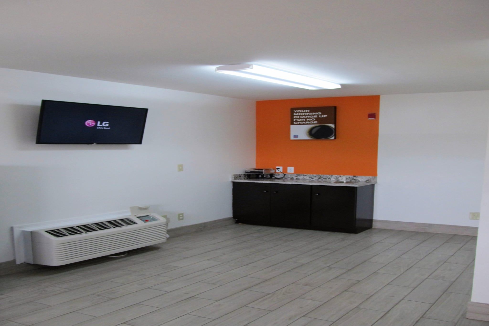 A room with a flat-screen TV mounted on the wall, a small kitchenette with a microwave, and a portable air conditioner.