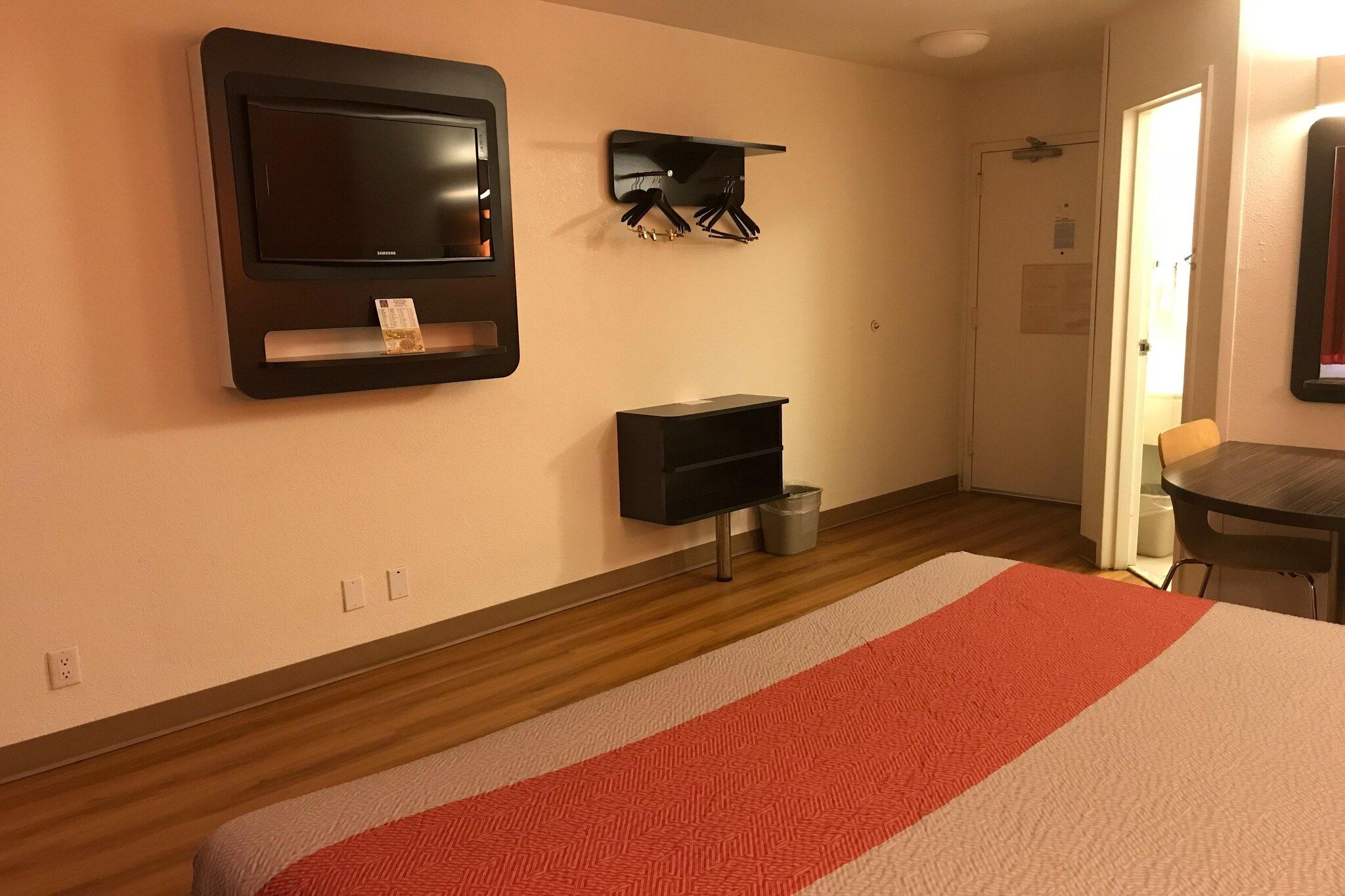A hotel room with a bed, a TV mounted on the wall, a coat rack, and a small table.
