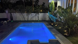 En rektangulär swimmingpool med en blå, upplyst vattenyta, omgiven av en vit vägg och olika tropiska växter.