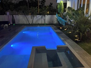 A rectangular swimming pool with a blue illuminated water surface, surrounded by a white wall and various tropical plants. - The Oasis Pool Villa (Chiang Mai)