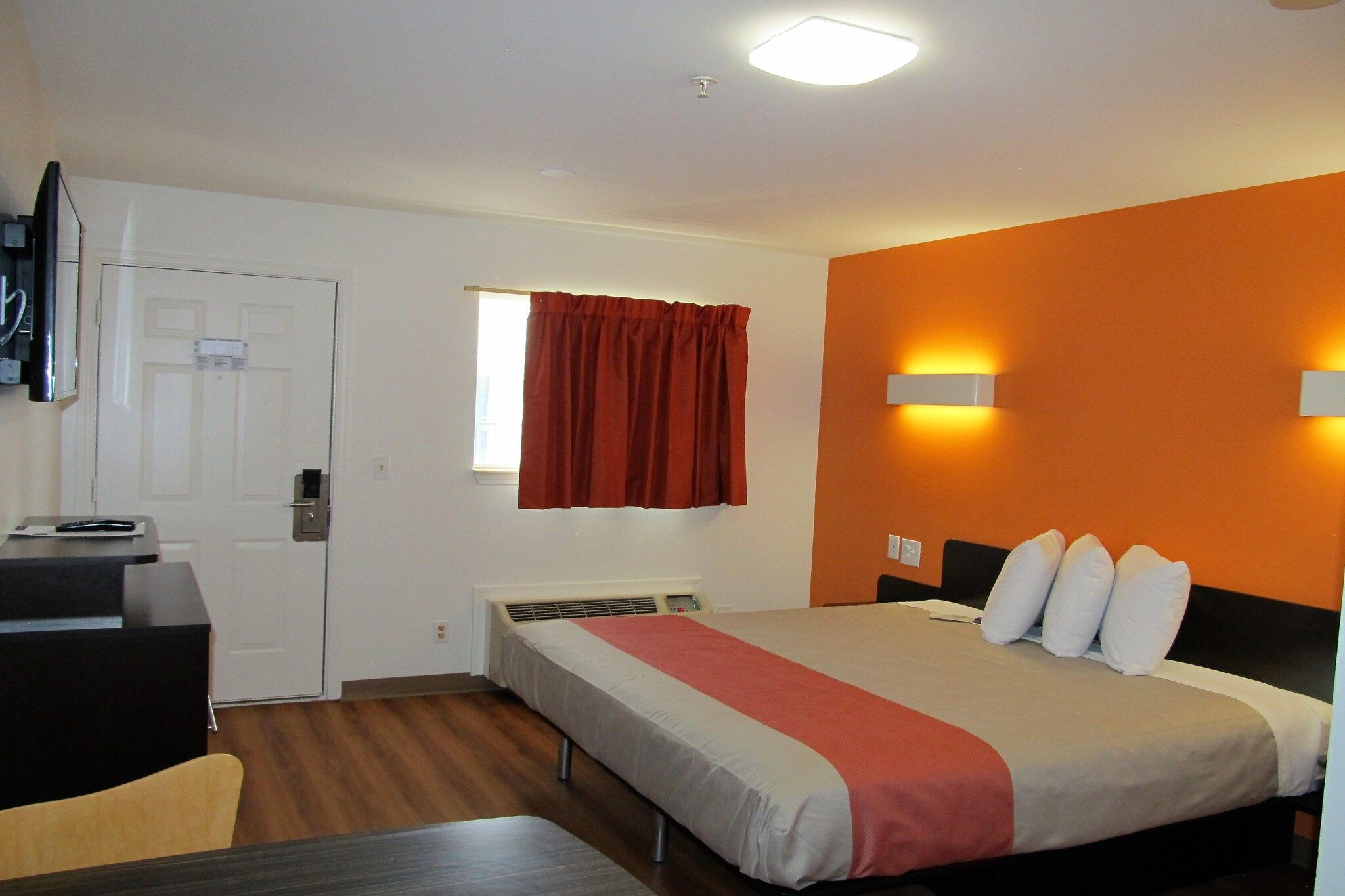 Habitación de hotel con cama, escritorio, silla, ventana con cortinas y lámpara de pared.