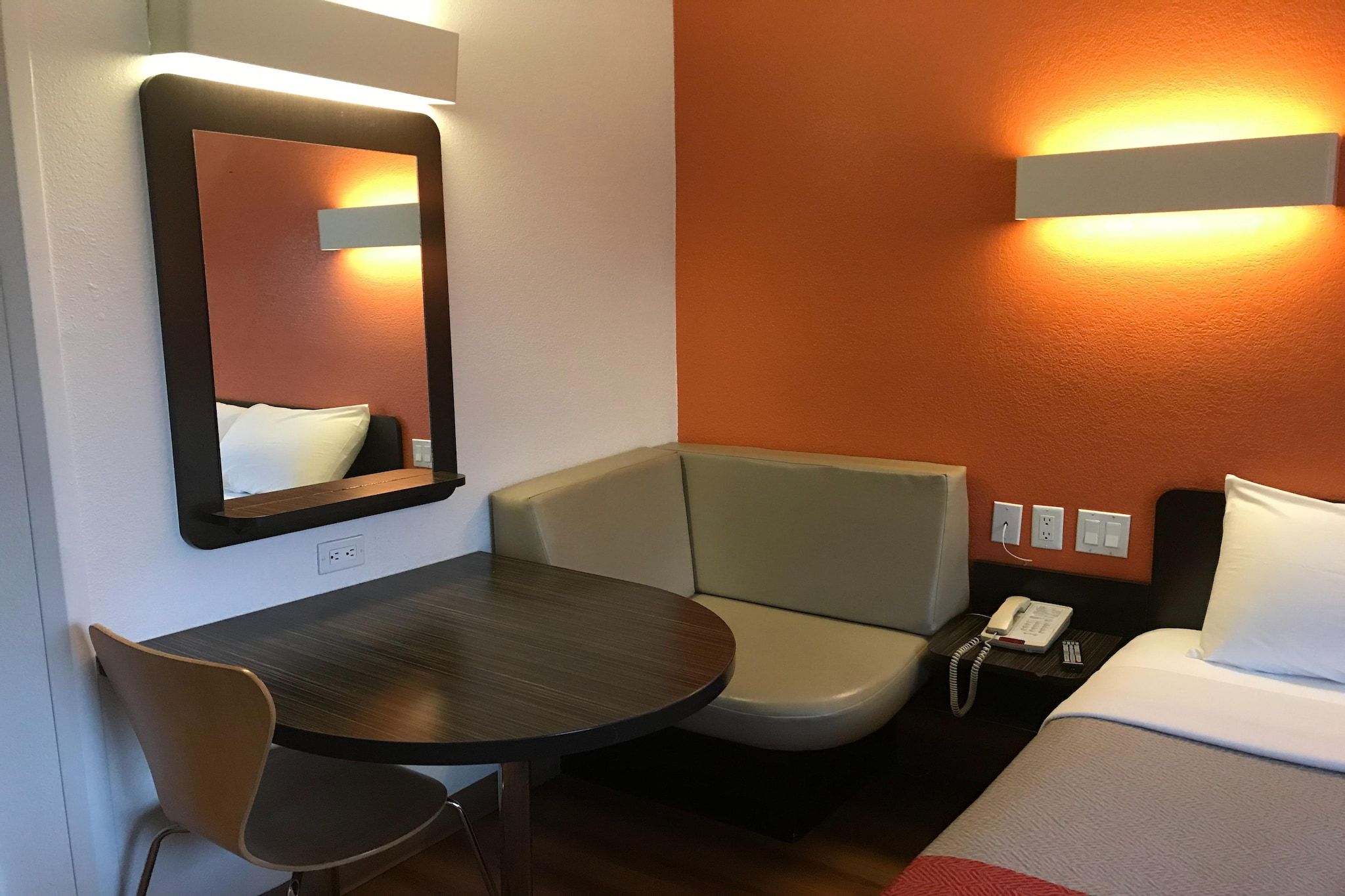 Ein Hotelzimmer mit Bett, kleinem Tisch, Stuhl, Nachttisch mit Telefon und einem an der Wand befestigten Spiegel.