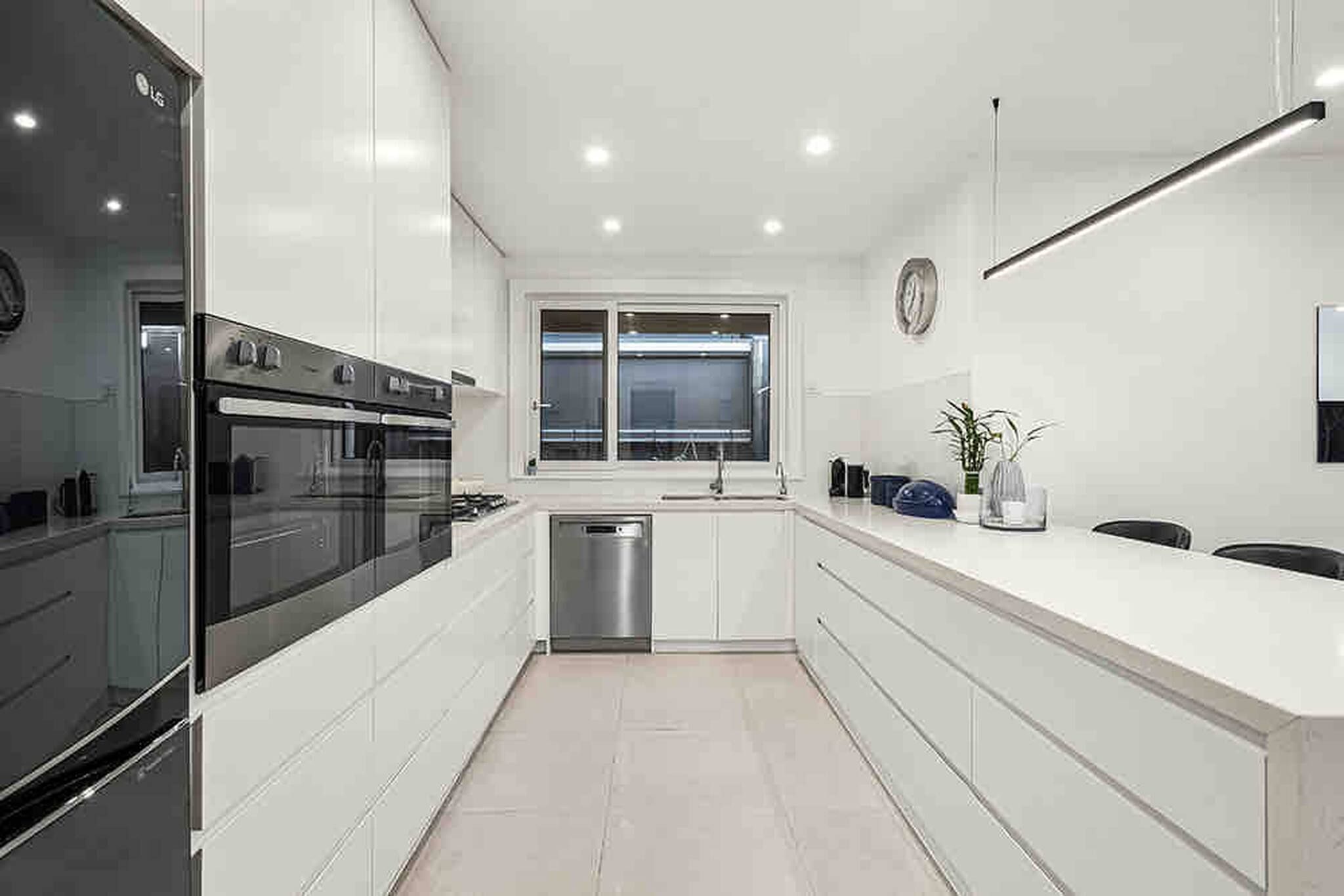 A modern kitchen with white cabinetry, a built-in oven, a stainless steel dishwasher, and a window above the sink.