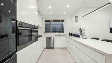 A modern kitchen with white cabinetry, a built-in oven, a stainless steel dishwasher, and a window above the sink.