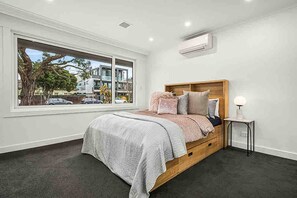 Un dormitorio con un ventanal grande, una cama con cabecera de madera, una mesita de noche y un aire acondicionado instalado en la pared.