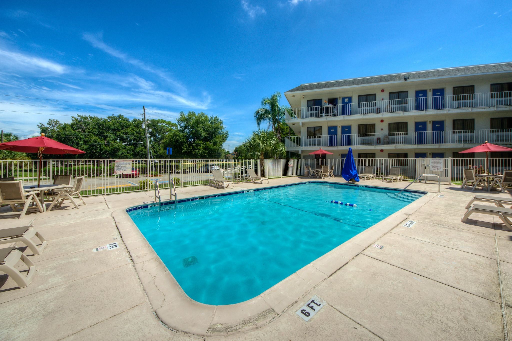 A swimming pool with lounge chairs and umbrellas, surrounded by a fence, with a building in the background.