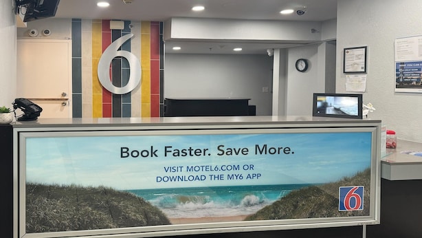 A reception desk with a large sign advertising a booking app, a number '6' sign, and a clock on the wall. - Cozy 1 Queen Bed | Mini Fridge (Bradenton)