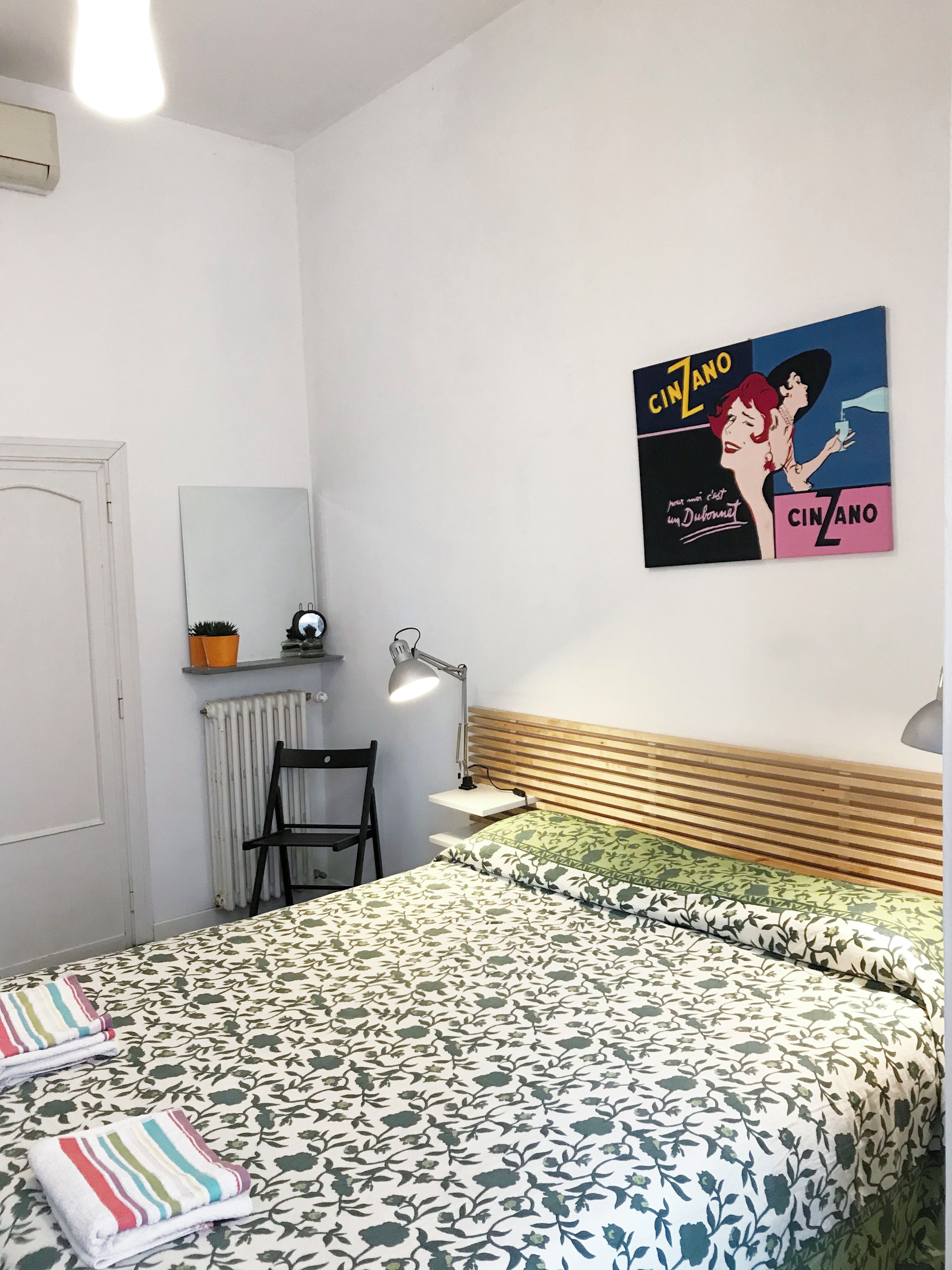 A bedroom with a bed, a chair, a desk, a lamp, a radiator, and a poster on the wall.