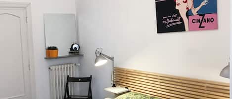 A bedroom with a bed, a chair, a desk, a lamp, a radiator, and a poster on the wall.
