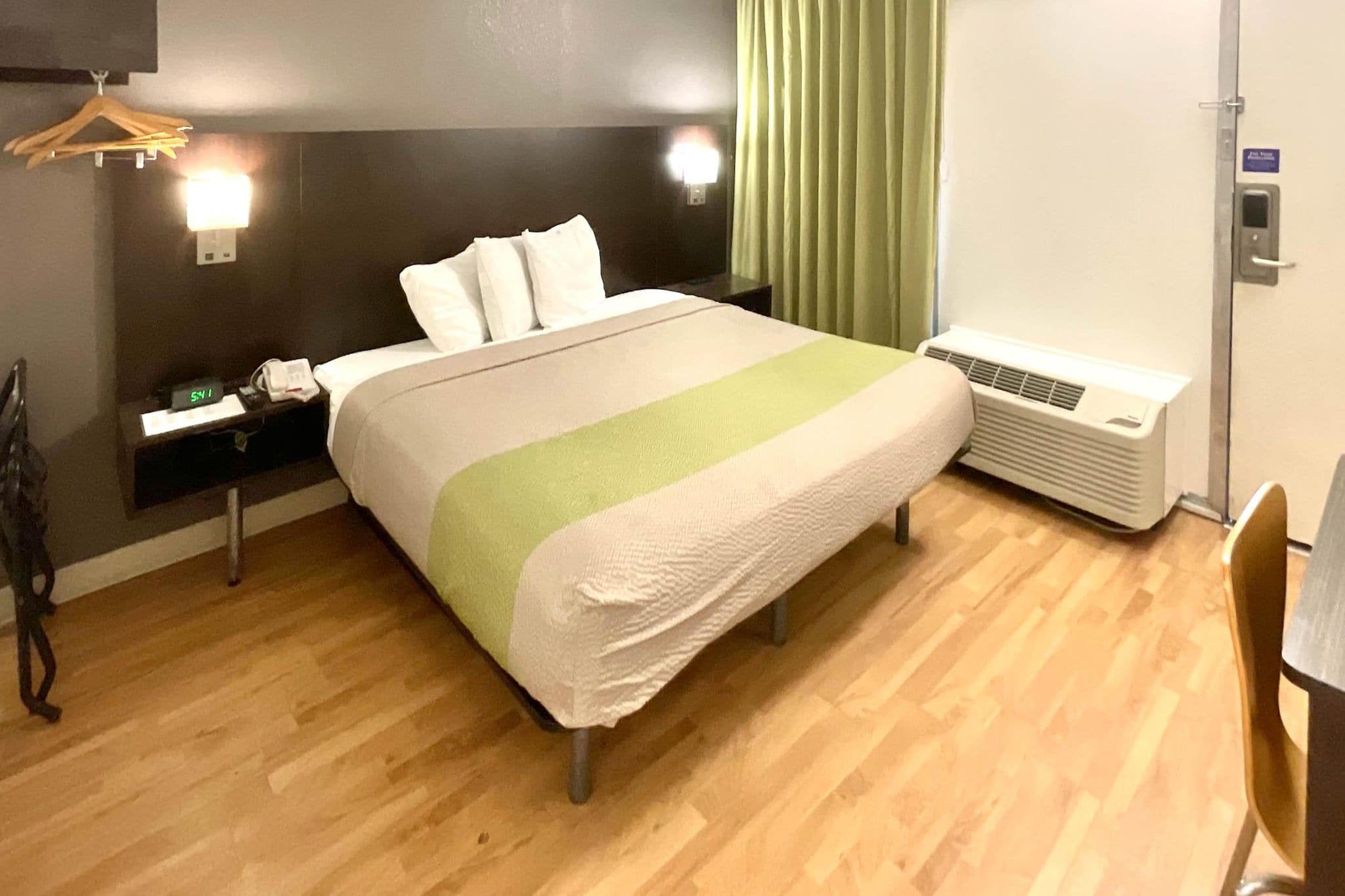 A hotel room with a bed, bedside table, lamp, air conditioning unit, and a chair.