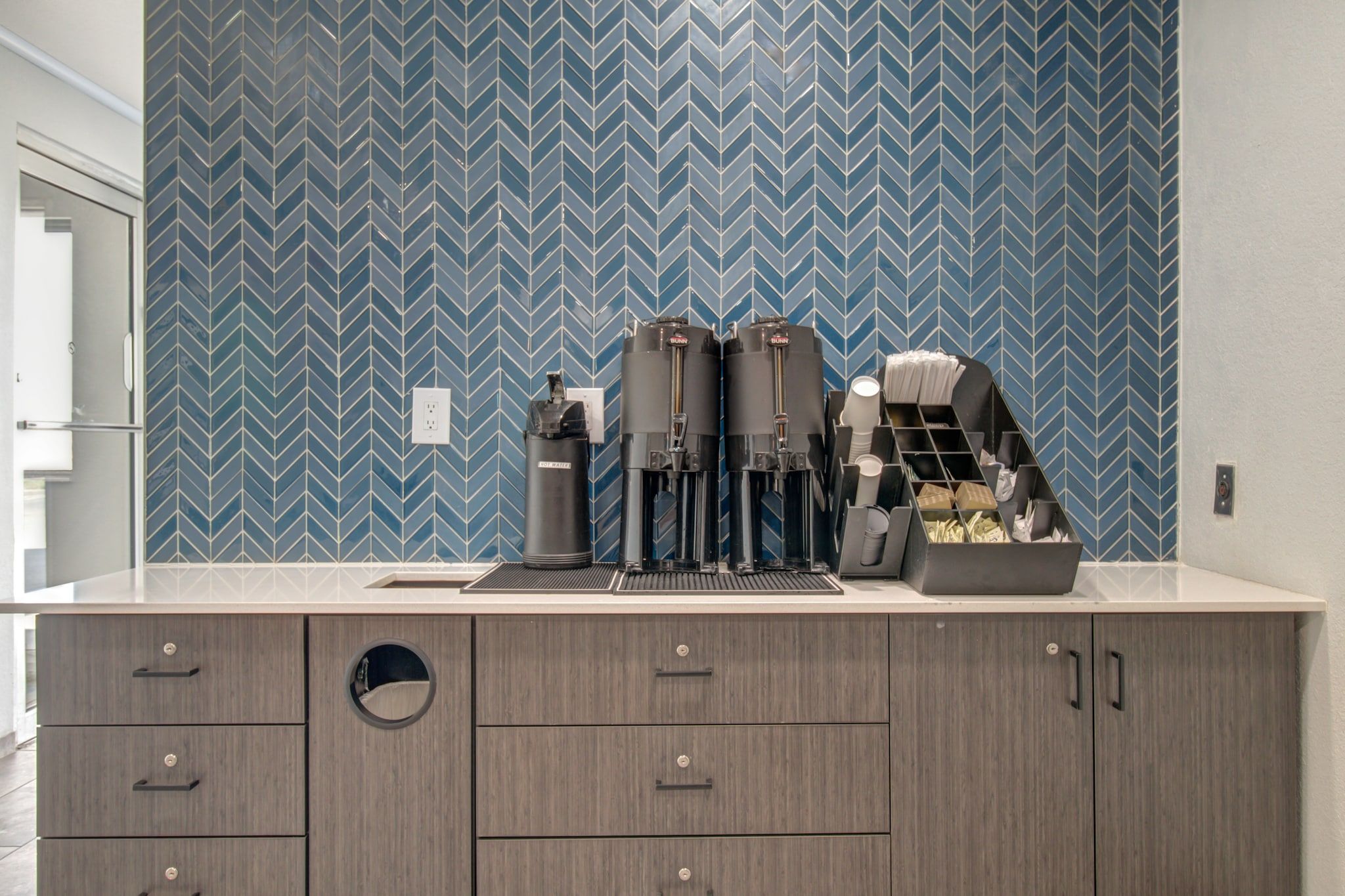 Een kitchenette met een blauwe backsplash van visgraat tegels, een koffieapparaat, een waterkoker en een koffieautomaat.