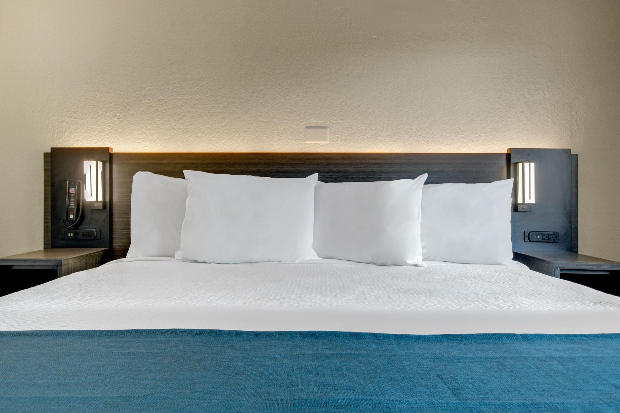 Een hotelkamer met een groot bed, witte beddengoed en een blauwe plaids. Twee nachtkastjes met lampen en een aan de muur bevestigd telefoon.