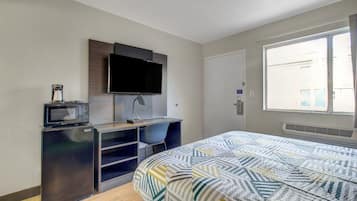 A bedroom with a bed, a desk with a computer, a television, and a window with a view.