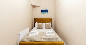 A narrow hotel room with a bed, pillows, a radiator, and a small picture on the wall. - South Kensington Base Near Knightsbridge (London)