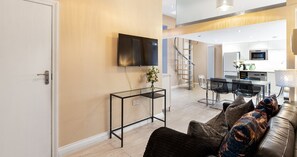 A modern living room with a sofa, a glass coffee table, a TV mounted on the wall, and a staircase leading to the upper floor. - South Kensington Base Near Knightsbridge (London)