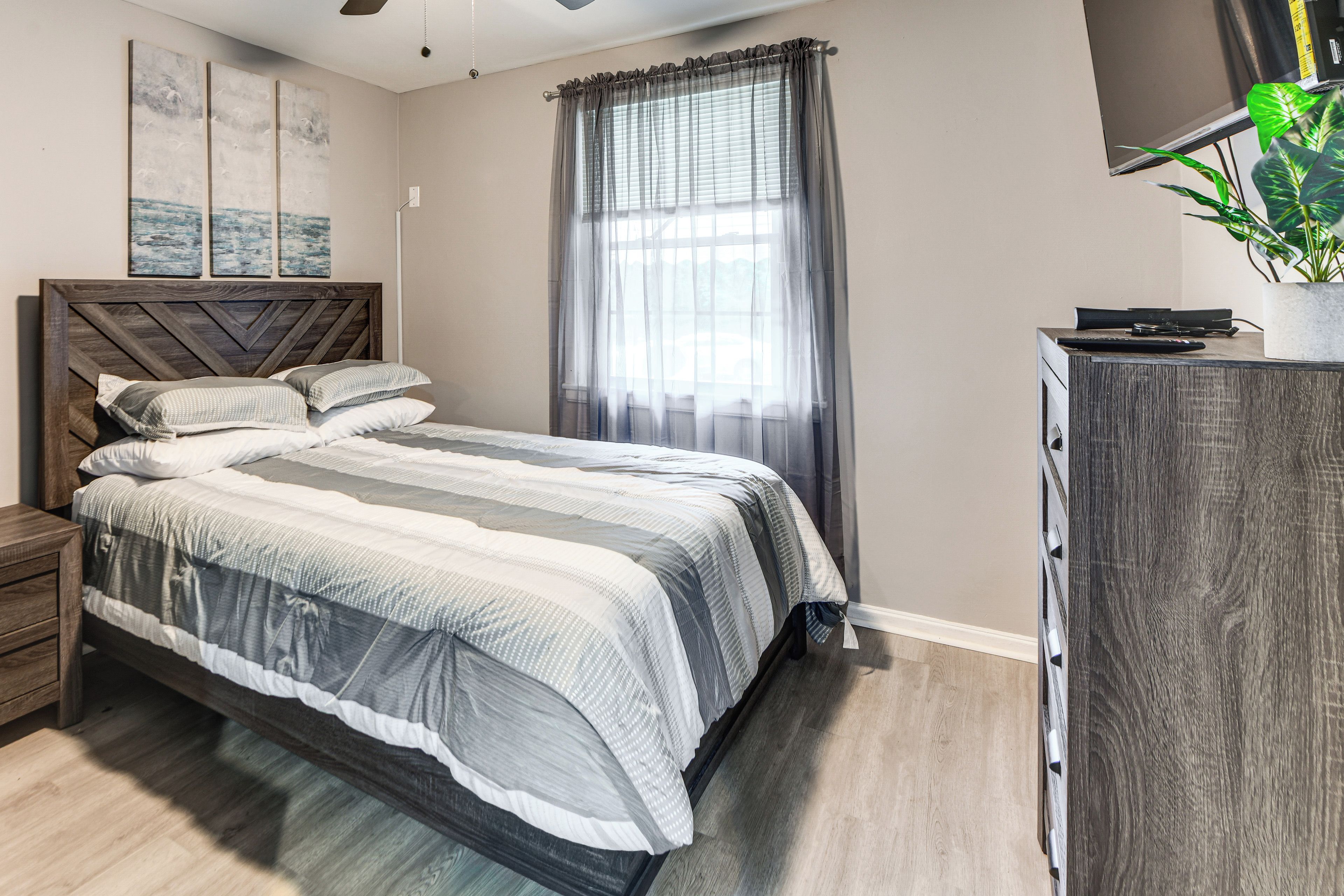 A bedroom with a bed, a nightstand, a dresser, a television, and a window with curtains.