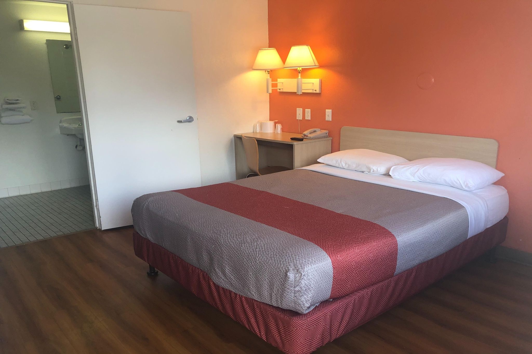 A hotel room with a bed, a desk with a lamp, and a bathroom visible through an open door.