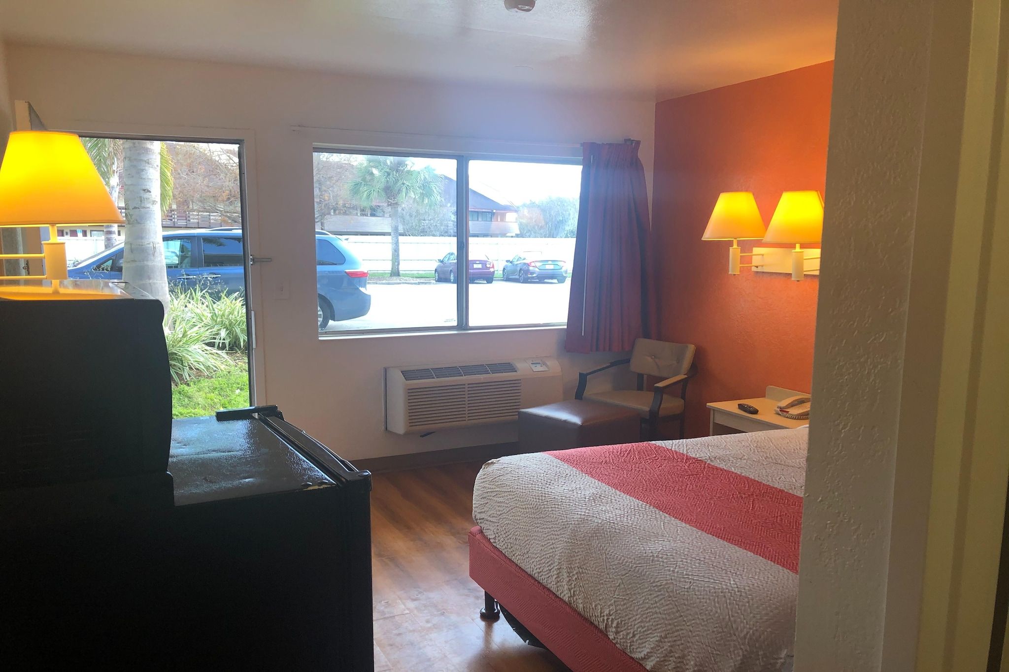 A hotel room with a bed, a desk, a chair, a heater, and a window with a view of parked cars and trees.