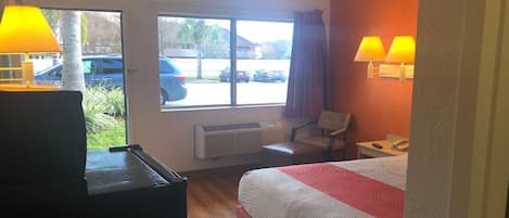 Habitación de hotel con cama, escritorio, silla, calefactor y ventana que ofrece vistas a coches aparcados y árboles.