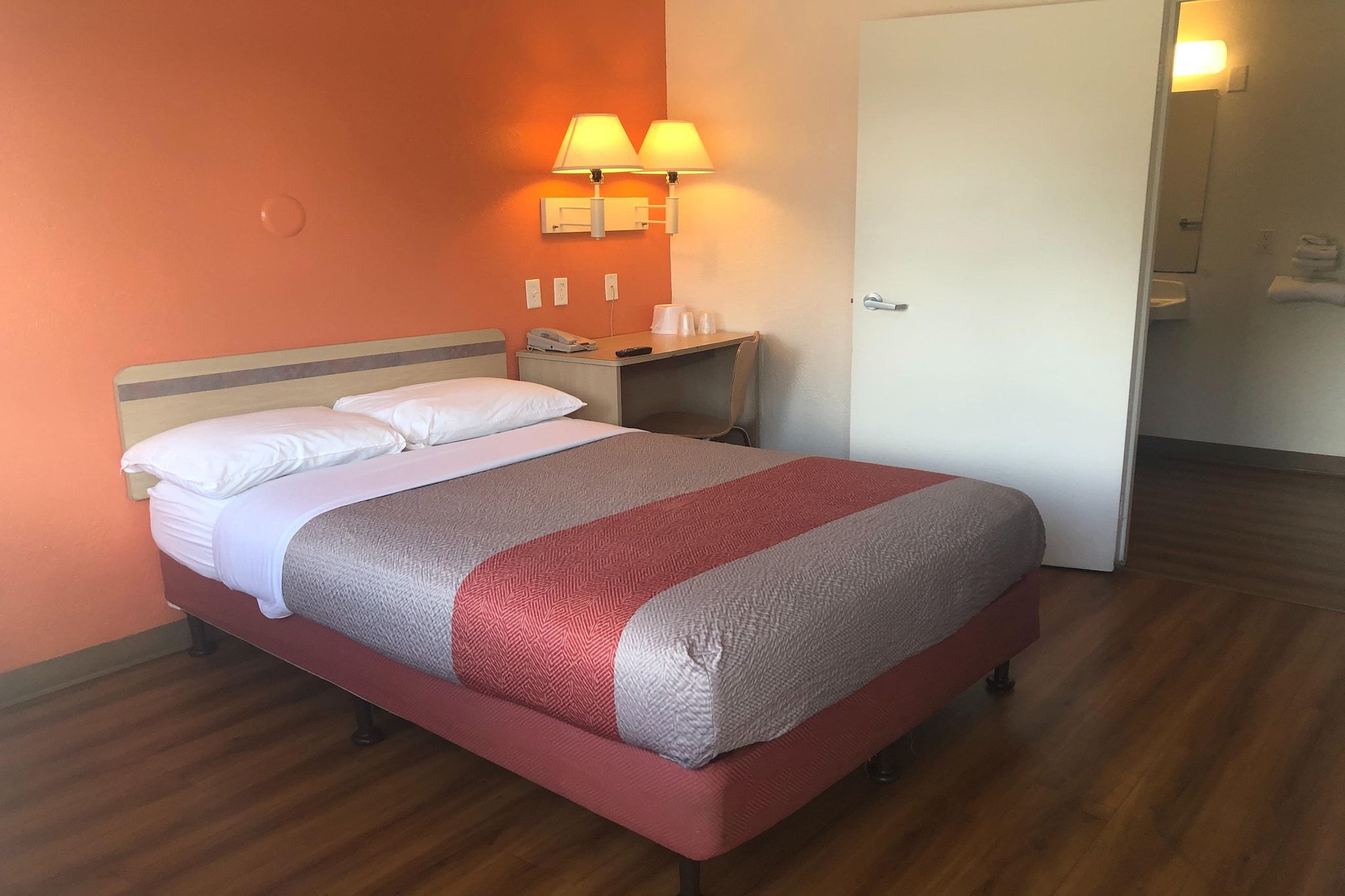 A hotel room with a bed, a nightstand, two wall-mounted lamps, a door, and a wooden floor.