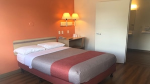 Quarto de hotel com cama, mesa de cabeceira, duas luminárias na parede, porta e piso de madeira.