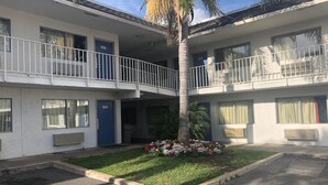 Um motel de dois andares com portas numeradas, um pátio central com uma palmeira e um pequeno canteiro de flores.