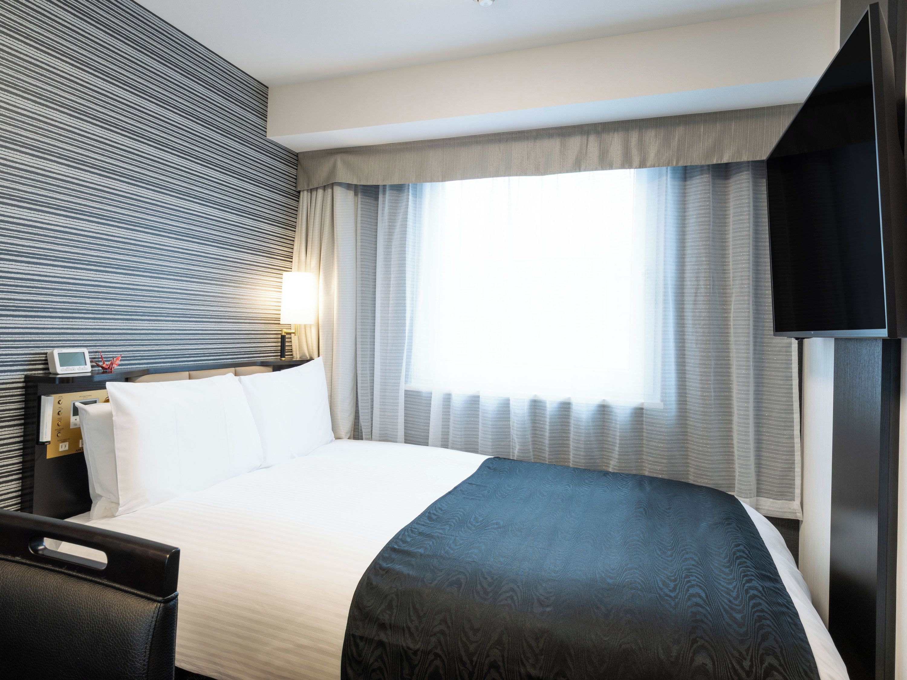 A hotel room with a bed, a bedside table with a lamp, a television, and a window with curtains.