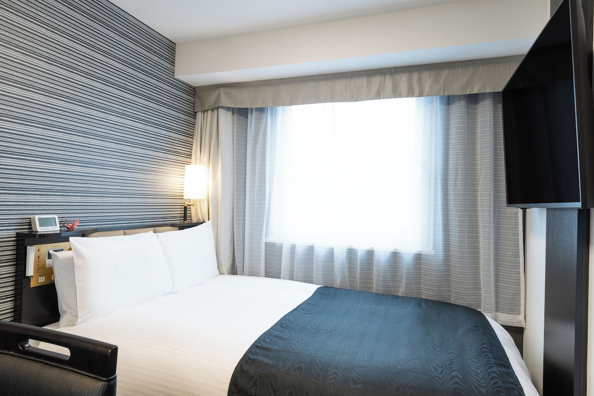 Ein Hotelzimmer mit Bett, Nachttisch mit Lampe, Fernseher und Fenster mit Vorhängen.