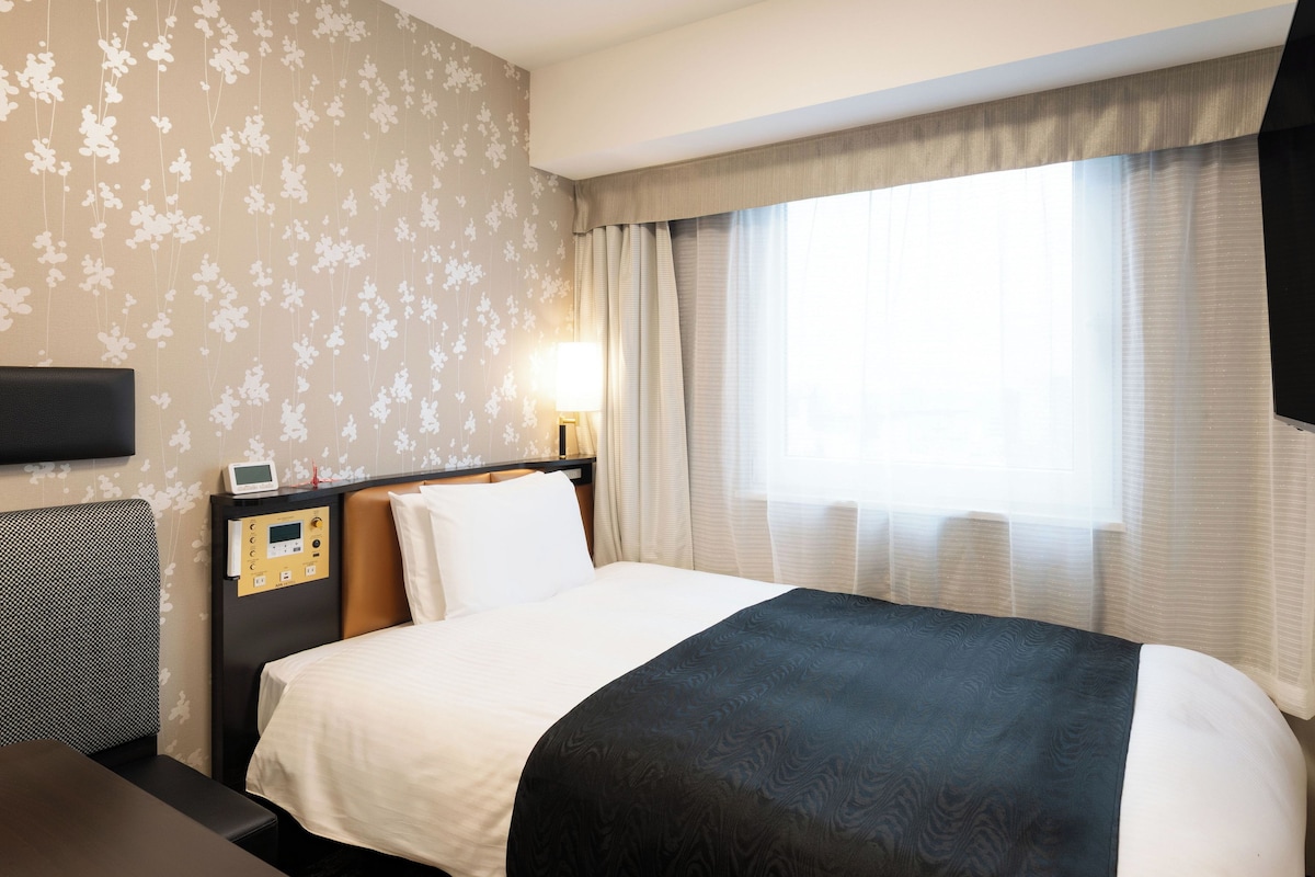 Ein Hotelzimmer mit Bett, Nachttisch mit Lampe, Wandfernseher und einem Fenster mit Vorhängen.