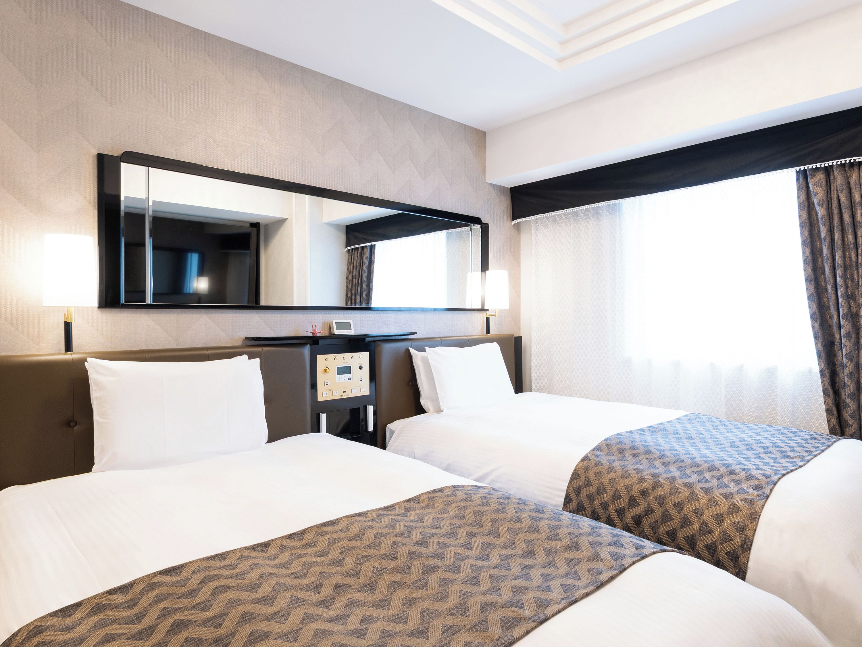 A hotel room with two beds, a bedside table, a mirror, and a window with curtains.