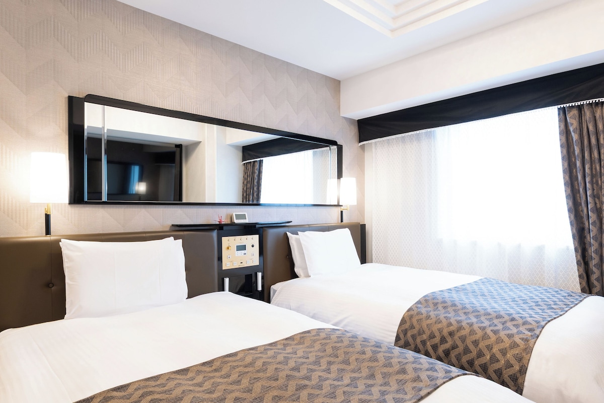 Ein Hotelzimmer mit zwei Betten, einem Nachttisch, einem Spiegel und einem Fenster mit Vorhängen.