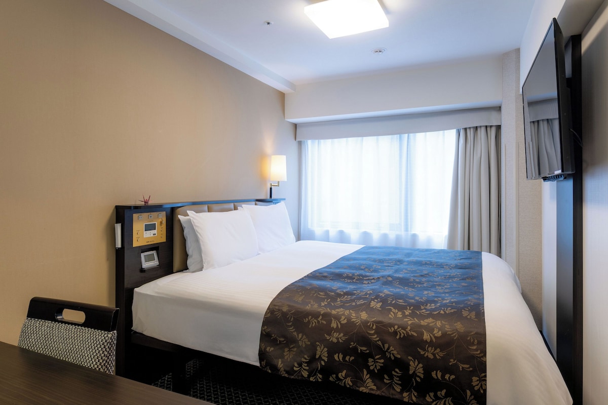 Ein Hotelzimmer mit einem Bett, Nachttisch, Lampe und einem Fenster mit Vorhängen.