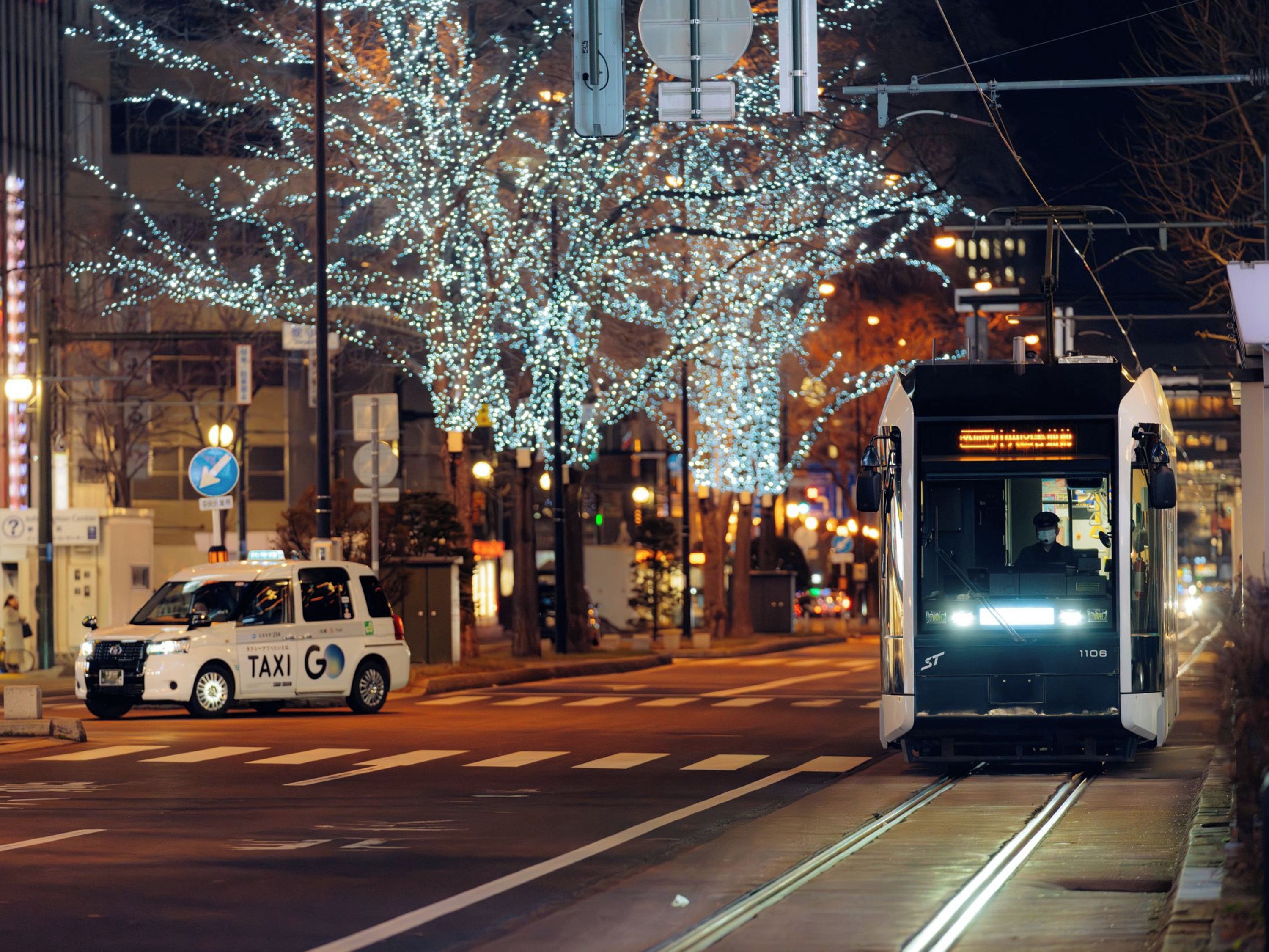 A tram on tracks with a white taxi in front, a tree with lights, and a building with signage.