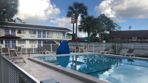 Una piscina con sillas de descanso y sombrillas, rodeada por una valla, con un edificio al fondo.