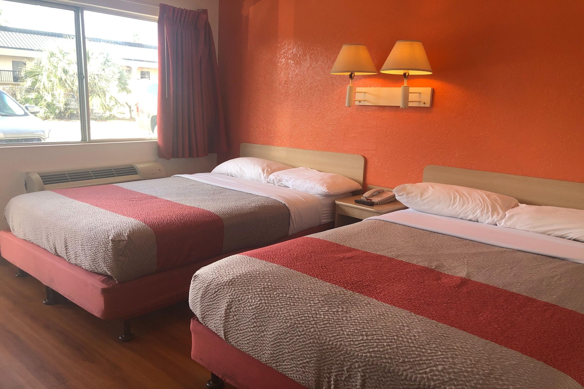 Une chambre d’hôtel avec deux lits, une fenêtre avec des rideaux, une lampe fixée au mur et un appareil de climatisation visible.