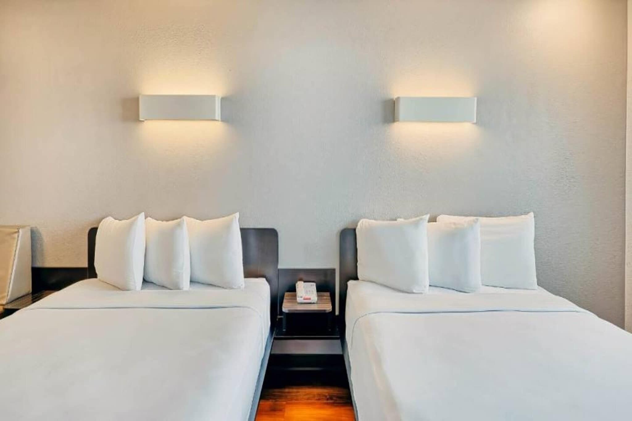 Een hotelkamer met twee bedden, elk met witte beddengoed en kussens, een klein nachtkastje met een lamp, en wandlampen.