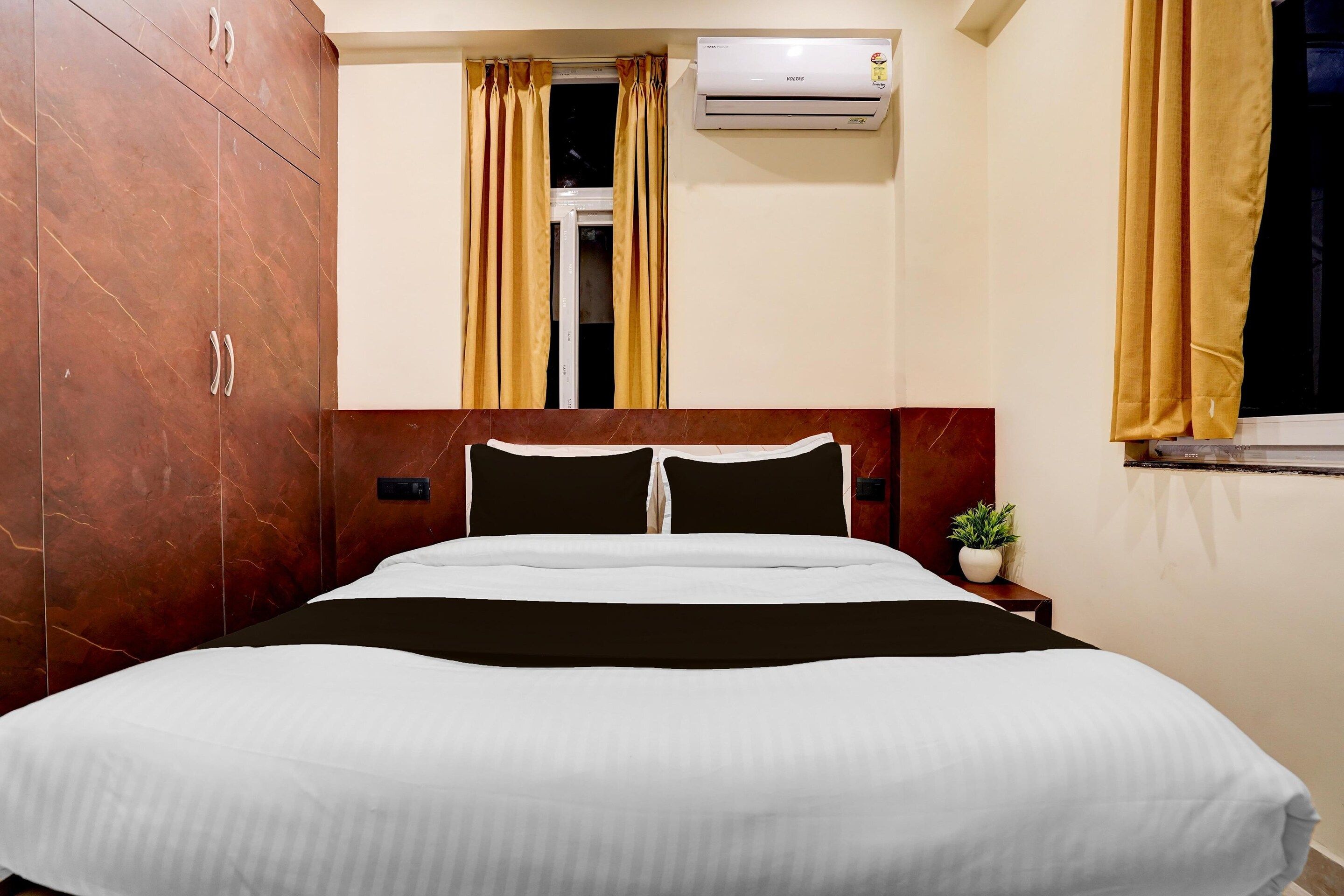 A hotel room with a bed, a nightstand, an air conditioning unit, a window with curtains, and a wooden wardrobe.
