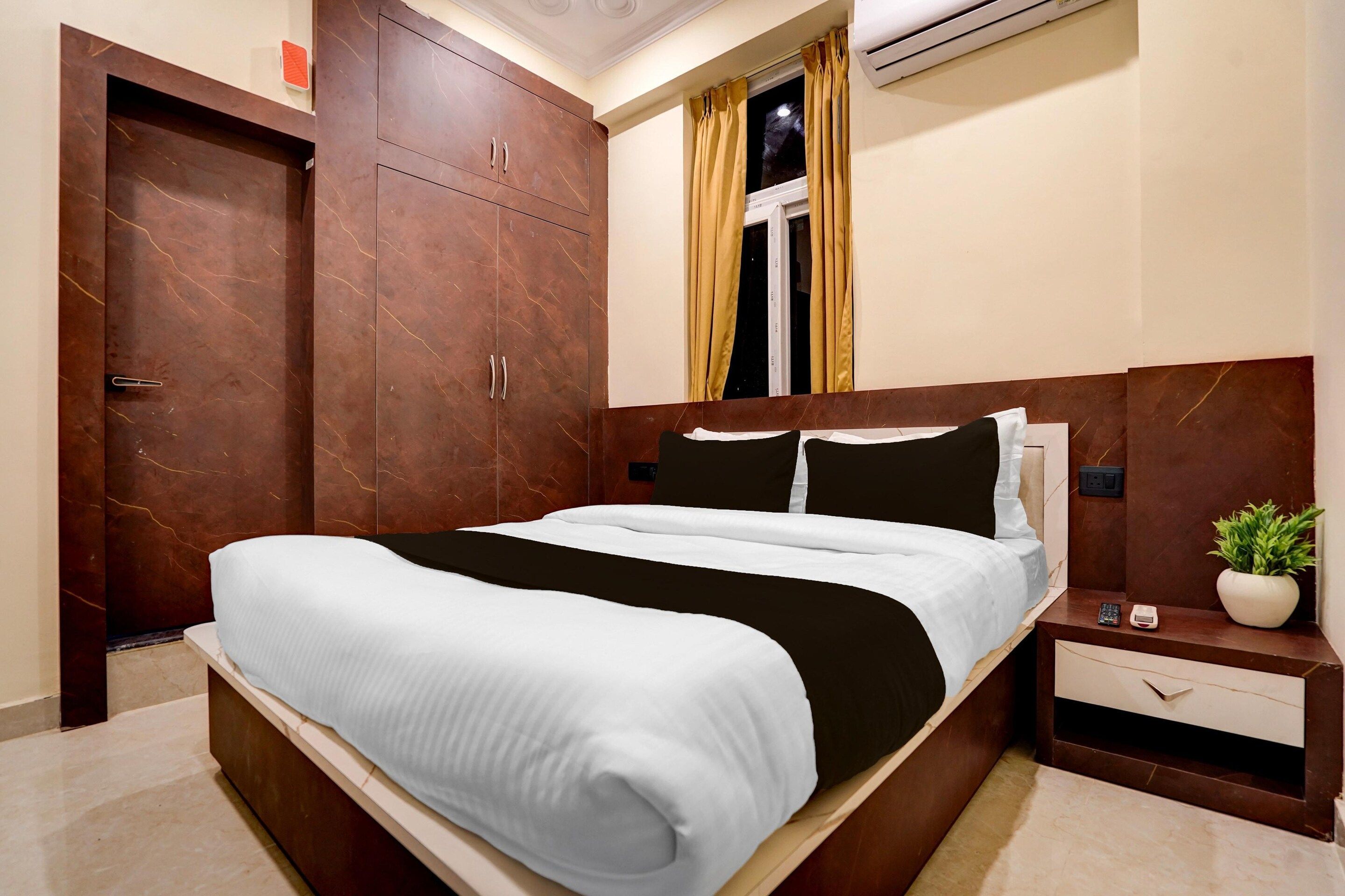 A bedroom with a bed, a wardrobe, a nightstand, a potted plant, and a window with curtains.