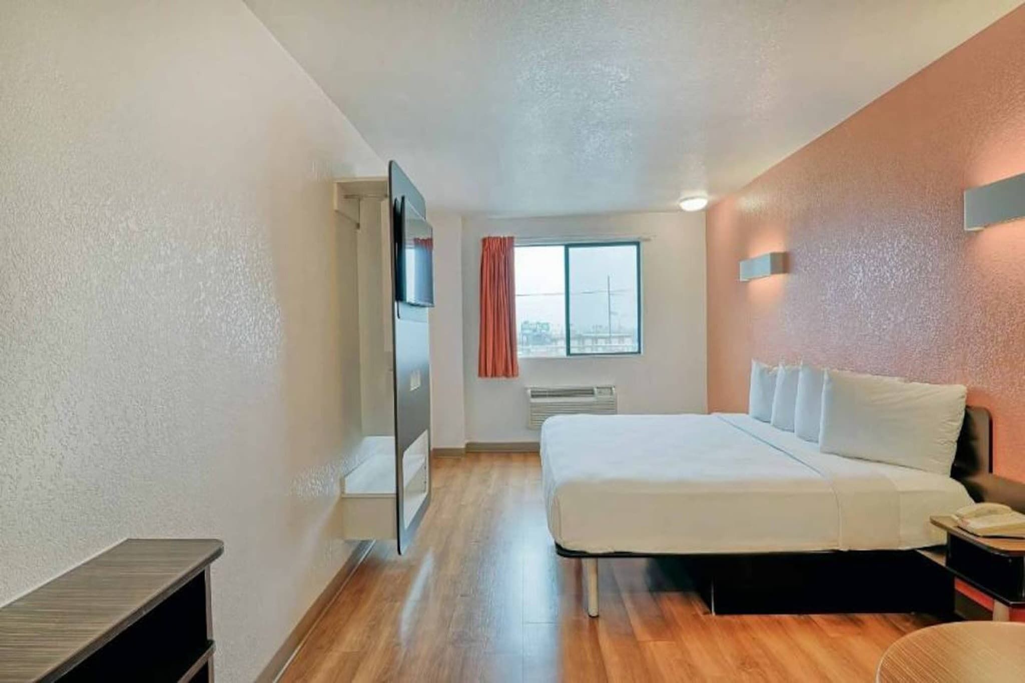 A hotel room with a bed, a nightstand, a window with curtains, a radiator, and a door leading to another room.