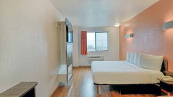 A hotel room with a bed, a nightstand, a window with curtains, a radiator, and a door leading to another room.