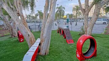 Een park met kunstbomen, een omheind gebied en een speeltuin met rode en grijze speeltoestellen.