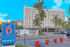 A motel with a large building, a sign indicating the entrance, and a parking lot with red planters. - 1 King Bed|Mini Fridge (Inglewood)