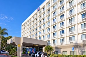 Et hotel med flere etager, en flad facade, en overdækket indgang og en parkeringsplads med biler.